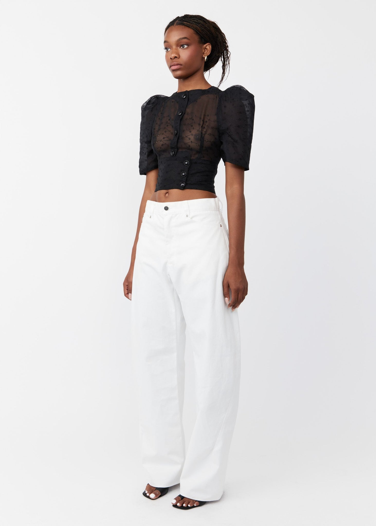 Model wearing a black sheer top and white pants on a white background